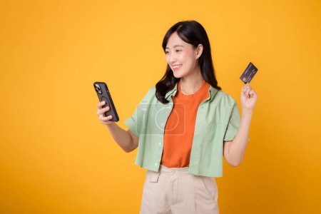Foto de Experimente la emoción de las compras móviles con una joven mujer asiática vibrante de 30 años, vistiendo una camisa naranja y un jersey verde, utilizando un teléfono inteligente mientras presenta la tarjeta de crédito en el fondo amarillo del estudio. - Imagen libre de derechos