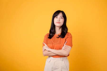 Téléchargez les photos : Libérez le pouvoir de la confiance alors qu'une jeune femme asiatique trentaine, revêtue d'une chemise orange, montre un geste de signe du bras croisé sur fond jaune. - en image libre de droit
