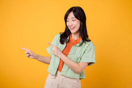 Photo for Young 30s Asian woman wearing green shirt on orange shirt, pointing fingers to free copy space with enthusiasm. concept of discount shopping promotion and discover exciting deals in image. - Royalty Free Image
