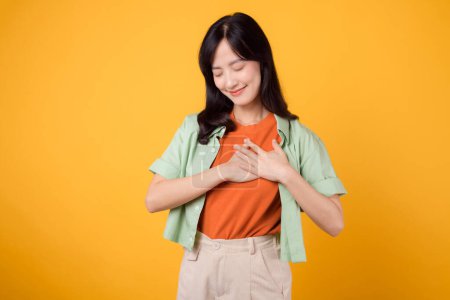 Téléchargez les photos : Bien-être comme une femme asiatique trentaine, portant une chemise verte, tient sa main sur sa poitrine sur une toile de fond orange. Concept de soins de santé sur fond jaune vif. - en image libre de droit