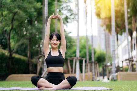 Foto de Retrato en forma joven mujer asiática con ropa deportiva negro escuchar música relajante mientras se estira el músculo antes de hacer ejercicio de yoga en la estera de yoga en el parque natural verde. concepto de bienestar. - Imagen libre de derechos