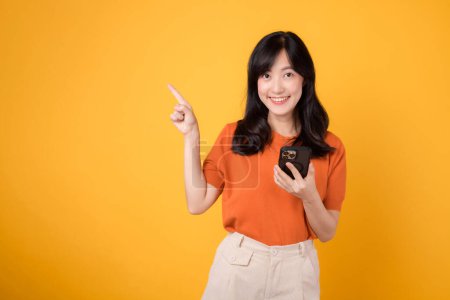 Téléchargez les photos : Jeune femme asiatique des années 30 portant une chemise orange pointant du doigt pour libérer de l'espace de copie tout en utilisant un smartphone isolé sur fond jaune. gaie femme avec nouveau concept de technologie d'application. - en image libre de droit