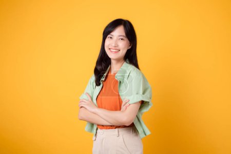 Photo for Confidence and well-being with a young 30s Asian woman wearing orange shirt. Her arm cross gesture on chest against a vibrant yellow background paints a portrait of self-assuredness and inner peace. - Royalty Free Image