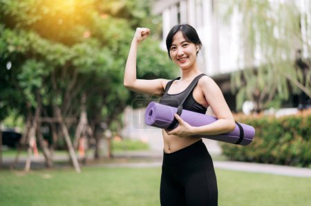 Photo for Embracing wellness: slim Asian girl prepped for a park workout, standing with rubber mat amidst greenery at sunset. Yoga and well-being. - Royalty Free Image