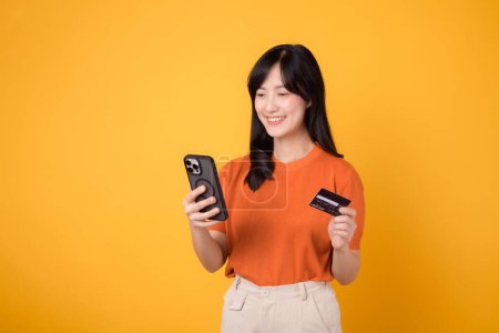 Photo for Smiling Asian woman in her 30s in orange shirt, using smartphone and holding credit card on yellow background. Online shopping joy. - Royalty Free Image