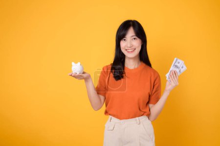 Photo for Cheerful Asian woman 30s holding piggy bank and cash money dollars on vibrant yellow background. Financial wealth concept. - Royalty Free Image