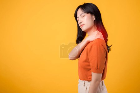 Téléchargez les photos : Femme asiatique bienveillante trentaine, portant une chemise orange, tient son épaule douloureuse sur fond jaune. Thérapie des maux de cou concept de syndrome de bureau médical. - en image libre de droit
