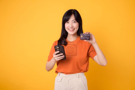 Photo for Enthusiastic young Asian woman 30s, using smartphone and holding credit card on yellow background. Swift online shopping. - Royalty Free Image