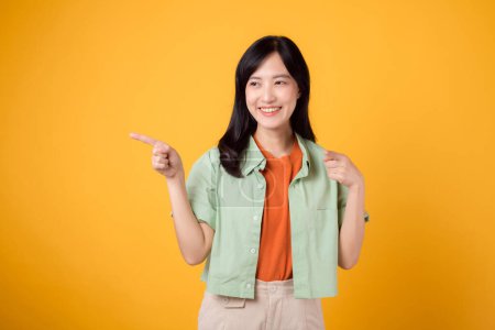 Téléchargez les photos : 30s jeune femme asiatique portant une chemise verte sur une chemise orange, pointant avec confiance les doigts pour libérer de l'espace de copie. Explorez le concept de promotion des achats à rabais avec une image attrayante. - en image libre de droit