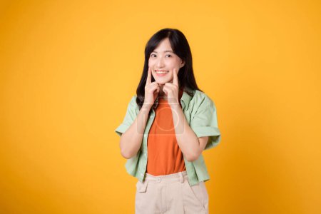 Téléchargez les photos : Jeune femme asiatique trentaine portant chemise orange et pull vert pointant vers ses dents isolées sur fond jaune. concept de soins dentaires. - en image libre de droit