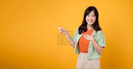 Photo for Young Asian woman 30s wearing green on orange shirt energetically points fingers to free copy space while holding her chest isolated on yellow background. discount shopping promotion concept. - Royalty Free Image
