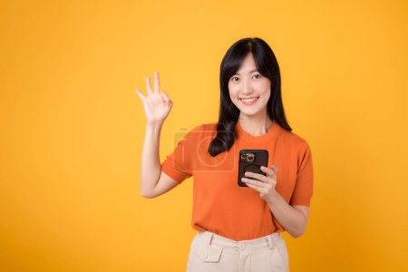 Téléchargez les photos : Joyeuse jeune femme asiatique trentenaire, vêtue d'une chemise orange, utilisant un smartphone avec un signe correct sur fond jaune vif. Nouveau concept d'application mobile - en image libre de droit