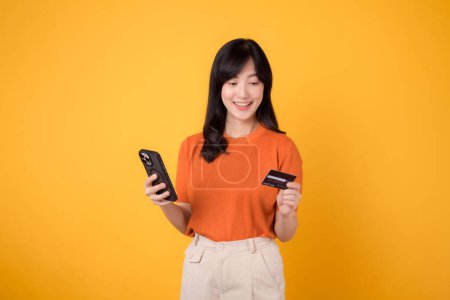 Photo for Happy young Asian woman in her 30s in orange shirt, using smartphone and holding credit card on yellow background. Seamless online shopping. - Royalty Free Image