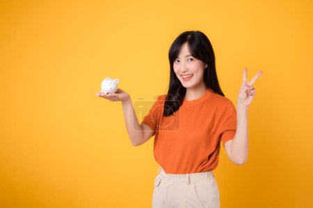 Photo for Smiling Asian woman in her 30s holding piggy bank, showing two hand sign, on vibrant yellow background. Financial growth concept. - Royalty Free Image