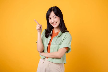 Photo for Unlock savings and shop now! young Asian woman 30s, wearing a green shirt on an orange shirt. happy face and pointing finger to free copy space against vibrant yellow backdrop. - Royalty Free Image