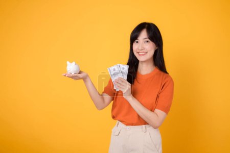 Photo for Smiling Asian woman 30s holding piggy bank and cash money dollars on vibrant yellow background. Wealth and savings concept. - Royalty Free Image