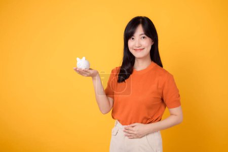 Photo for Happy Asian woman 30s with piggy bank, standing on vibrant yellow background. Wealth and saving concept. - Royalty Free Image