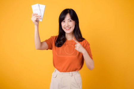 Photo for Radiant Asian woman 30s points to cash money dollars on vibrant yellow backdrop. Financial success concept. - Royalty Free Image