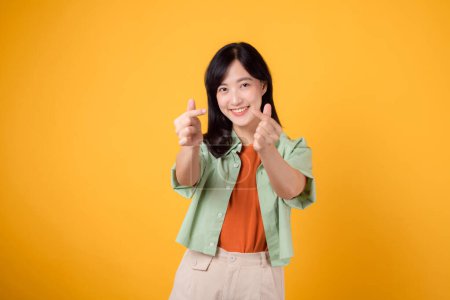 Téléchargez les photos : Bonheur avec une jeune femme asiatique trentenaire, vêtue d'une chemise orange et d'un pull vert. Son petit geste de cœur et son doux sourire expriment un message profond à travers le langage corporel. - en image libre de droit