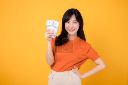 Photo for Energetic Asian woman 30s, holding cash money dollars, standing on vibrant yellow backdrop. - Royalty Free Image