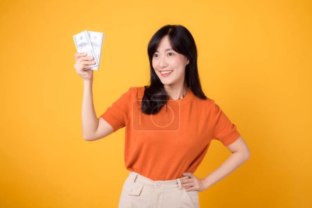 Photo for Vivacious young Asian woman 30s, holding cash money dollars, standing on vibrant yellow backdrop. - Royalty Free Image