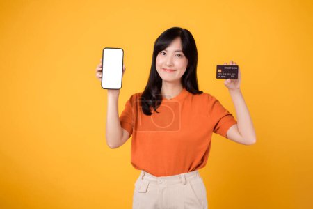Foto de Mujer asiática dinámica de 30 años, presentando smartphone y tarjeta de crédito sobre fondo amarillo. Compras de pago online sin esfuerzo. - Imagen libre de derechos