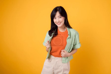 Photo for Joy of well-being with a cheerful young Asian woman 30s wearing an orange shirt. happy mind wellness gesture on a yellow background, capturing a happiness portrait of inner bliss. - Royalty Free Image