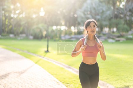 Foto de Una corredora. en forma joven mujer asiática de 30 años, con ropa deportiva rosa y sosteniendo una toalla, mientras corre en un parque público al atardecer. Acepta el concepto de bienestar y actividad al aire libre. - Imagen libre de derechos