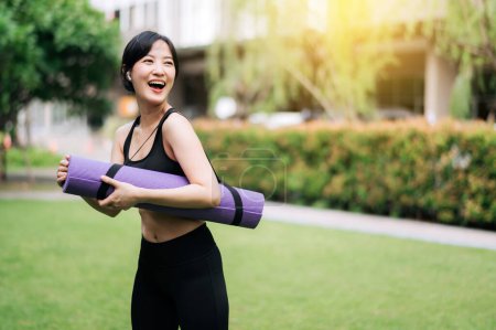 Téléchargez les photos : La sérénité au coucher du soleil rencontre le yoga : jeune femme asiatique, tapis en caoutchouc à la main, anticipe l'entraînement au parc. Santé, équilibre et bien-être naturel. - en image libre de droit