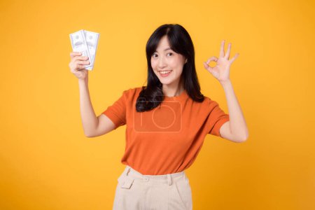 Photo for Positive young Asian woman 30s, pointing finger to cash money dollars, Okay hand sign, standing on vibrant yellow backdrop. - Royalty Free Image