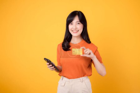 Photo for Fresh Asian woman 30s, using smartphone and presenting credit card on vibrant yellow background. Speedy online shopping payment transaction. - Royalty Free Image