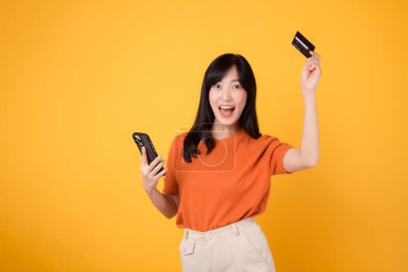 Téléchargez les photos : Contenu jeune femme asiatique dans la trentaine en chemise orange, en utilisant un smartphone et en tenant la carte de crédit sur fond jaune. Achats en ligne gratifiants. - en image libre de droit