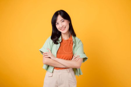 Photo for Confidence and well-being with a young 30s Asian woman wearing an orange shirt. Her arm cross gesture on her chest against a vibrant yellow background radiates self-assuredness and inner peace. - Royalty Free Image