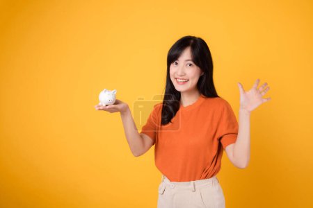 Photo for Smiling Asian woman in her 30s holding piggy bank, showing five hand sign, on vibrant yellow background. Financial growth concept. - Royalty Free Image