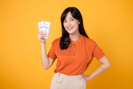 Photo for Radiant young Asian woman 30s, holding cash money dollars, standing on vibrant yellow backdrop. - Royalty Free Image