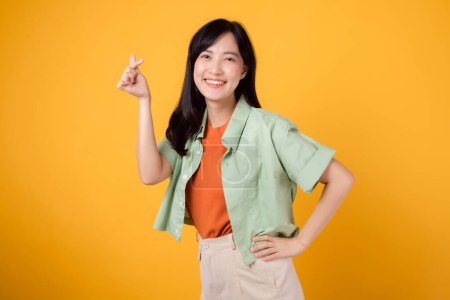 Photo for Happiness with a young Asian woman 30s, dressed in an orange shirt and green jumper. Her mini heart gesture, hand on hip, and gentle smile convey a profound message through body language. - Royalty Free Image