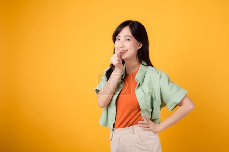 Foto de Abrace la felicidad con una joven mujer asiática de 30 años, vestida con una camisa naranja y un jersey verde. Su mini gesto del corazón, la sujeción de la cadera y su suave sonrisa transmiten un mensaje profundo a través del lenguaje corporal. - Imagen libre de derechos