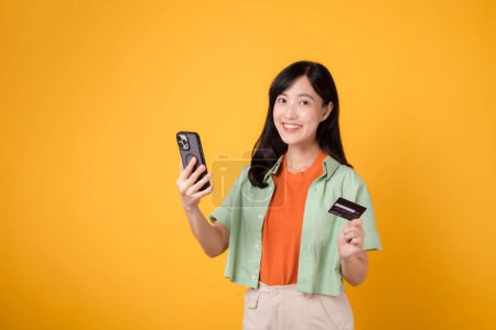 Foto de Compras desde el móvil con una cautivadora mujer asiática joven de 30 años, elegantemente vestida con camisa naranja y jersey verde, usando un teléfono inteligente mientras sostiene la tarjeta de crédito en el fondo del estudio amarillo. - Imagen libre de derechos