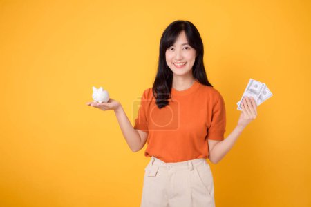 Photo for Dynamic Asian woman 30s holding piggy bank and cash money dollars on vibrant yellow background. Wealth and savings concept. - Royalty Free Image