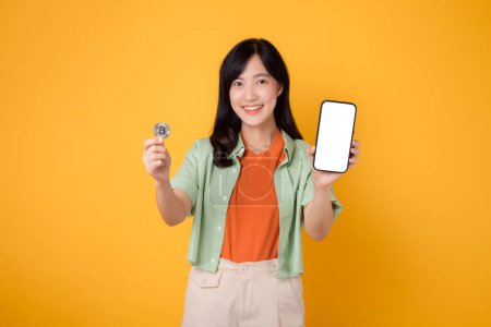 Téléchargez les photos : L'innovation de la finance future avec la jeune femme asiatique des années 30, élégamment vêtue d'une chemise orange et d'un pull vert, à l'aide d'un écran de smartphone et d'une pièce de monnaie cryptographique sur fond jaune. - en image libre de droit