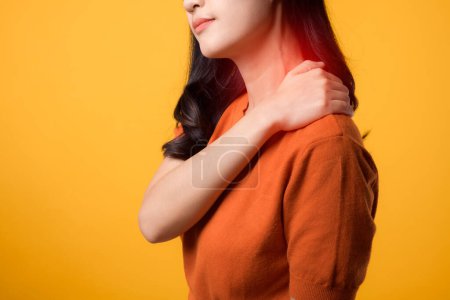 Foto de Mujer asiática de apoyo de 30 años, con una camisa naranja, sostiene su hombro dolorido sobre un fondo amarillo. Dolor de cuello terapia concepto de síndrome de consultorio médico. - Imagen libre de derechos