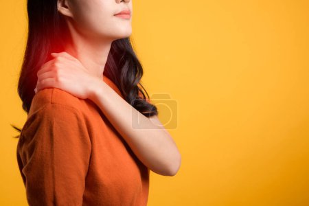 Photo for Aware young Asian woman 30s, wearing an orange shirt, holds her pain shoulder on yellow background. Neck ache therapy medical office syndrome concept. - Royalty Free Image