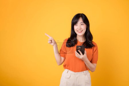 Téléchargez les photos : Embrassez le bonheur avec une jeune femme asiatique des années 30, portant une chemise orange, pointant vers l'espace libre de copie tout en utilisant un smartphone sur fond jaune. nouveau concept d'application. - en image libre de droit