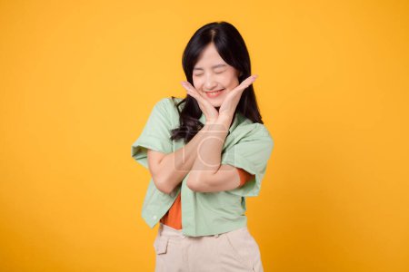 Foto de Una joven mujer asiática de 30 años que lleva una camisa verde en una camisa naranja irradia alegría con una cara feliz. Descubre la energía vibrante de la imagen alegre sobre un fondo amarillo. - Imagen libre de derechos