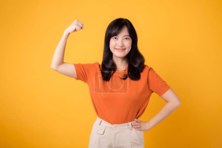 Téléchargez les photos : Confiante jeune femme asiatique trentaine célèbre avec un geste de signe de la main poing vers le haut, portant une chemise orange sur fond jaune. Concept d'autonomisation. Concept d'autonomisation et de féminisme. - en image libre de droit