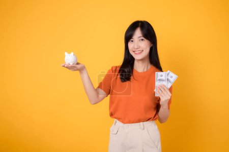 Téléchargez les photos : Femme asiatique rayonnante trentaine tenant tirelire et argent comptant dollars sur fond jaune vif. Concept de bien-être financier - en image libre de droit