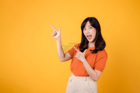 Téléchargez les photos : Répandre le bonheur avec une jeune femme asiatique trentaine, revêtant une chemise orange, pointant vers l'espace libre de copie sur fond jaune. recommander le concept de produit. - en image libre de droit