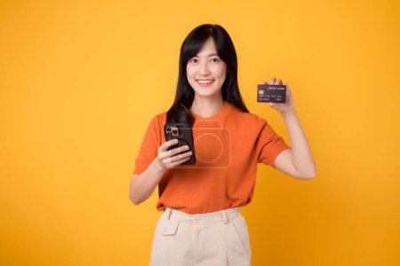 Téléchargez les photos : Vibrant jeune femme asiatique trentaine, en utilisant un smartphone et tenant la carte de crédit sur fond jaune. Achats en ligne pratiques. - en image libre de droit