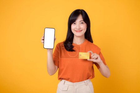 Foto de Mujer asiática entusiasta de 30 años, que muestra la pantalla del teléfono inteligente en blanco y la tarjeta de crédito sobre un fondo amarillo vibrante. Swift compras de pago en línea. - Imagen libre de derechos