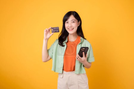 Foto de Descubre la alegría de las compras en línea con la alegre mujer asiática de 30 años, con camisa naranja y jersey verde, utilizando el teléfono inteligente para mostrar la tarjeta de crédito en el fondo del estudio amarillo. Concepto móvil. - Imagen libre de derechos